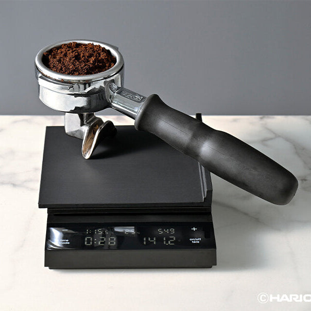 square silicone mat on top of a digital scale weighing a espresso portafilter with coffee ground. on a marble table and gray background.