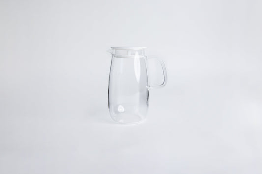 Round clear glass pitcher with glass handle and white lid set on a white background