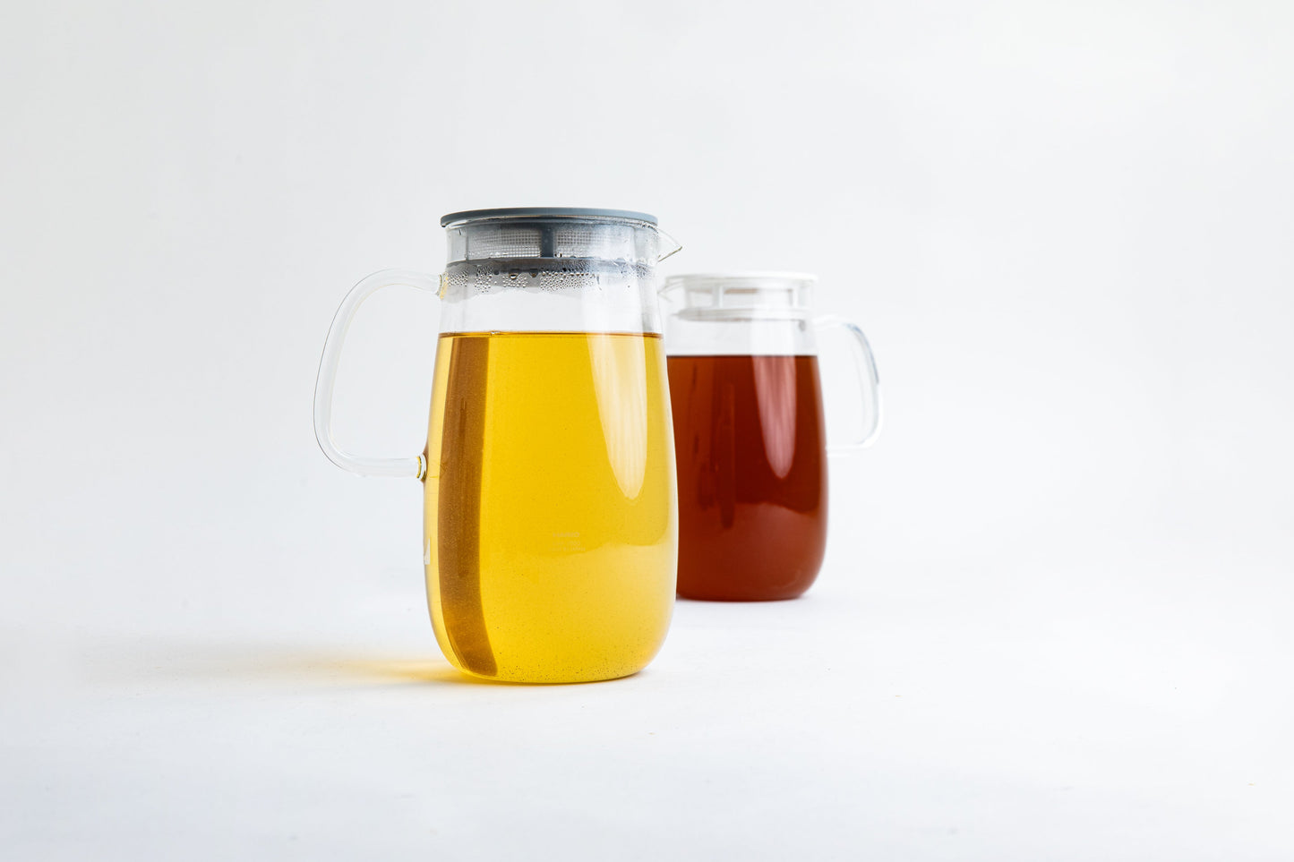 2 Round clear glass pitchers with glass handle and one has a white lid, and one with a gray lid. Gray lid pitcher is filled with a clear yellow liquid and white lid is filled with clear brown liquid set on a white background