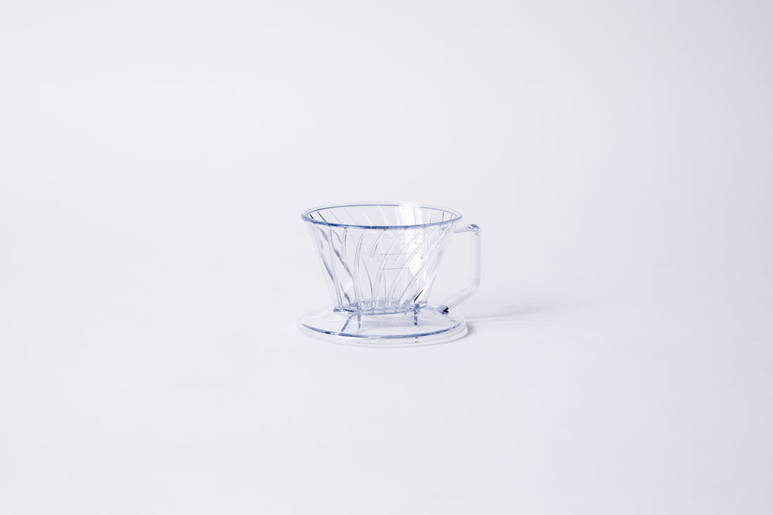 Clear plastic ribbed cone shaped dripper with a flat bottom base and handle. Set on a light gray background