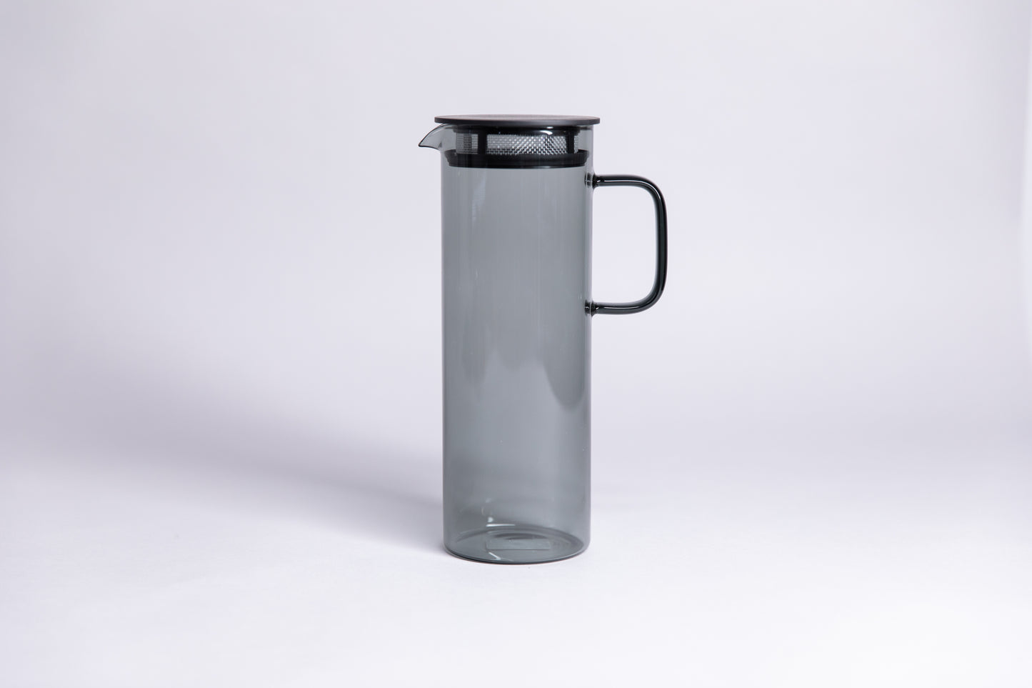 Cylindrical transparent gray colored glass pitcher with a fluted spout and rectangular handle with a black colored filtered lid. set on a a white background.