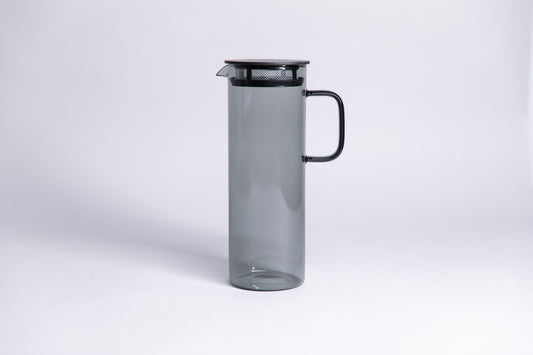 Cylindrical transparent gray colored glass pitcher with a fluted spout and rectangular handle with a black colored filtered lid. set on a a white background.