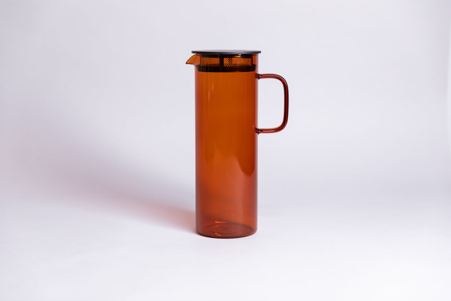 Cylindrical transparent amber colored glass pitcher with a fluted spout and rectangular handle with a black colored filtered lid. set on a a white background.