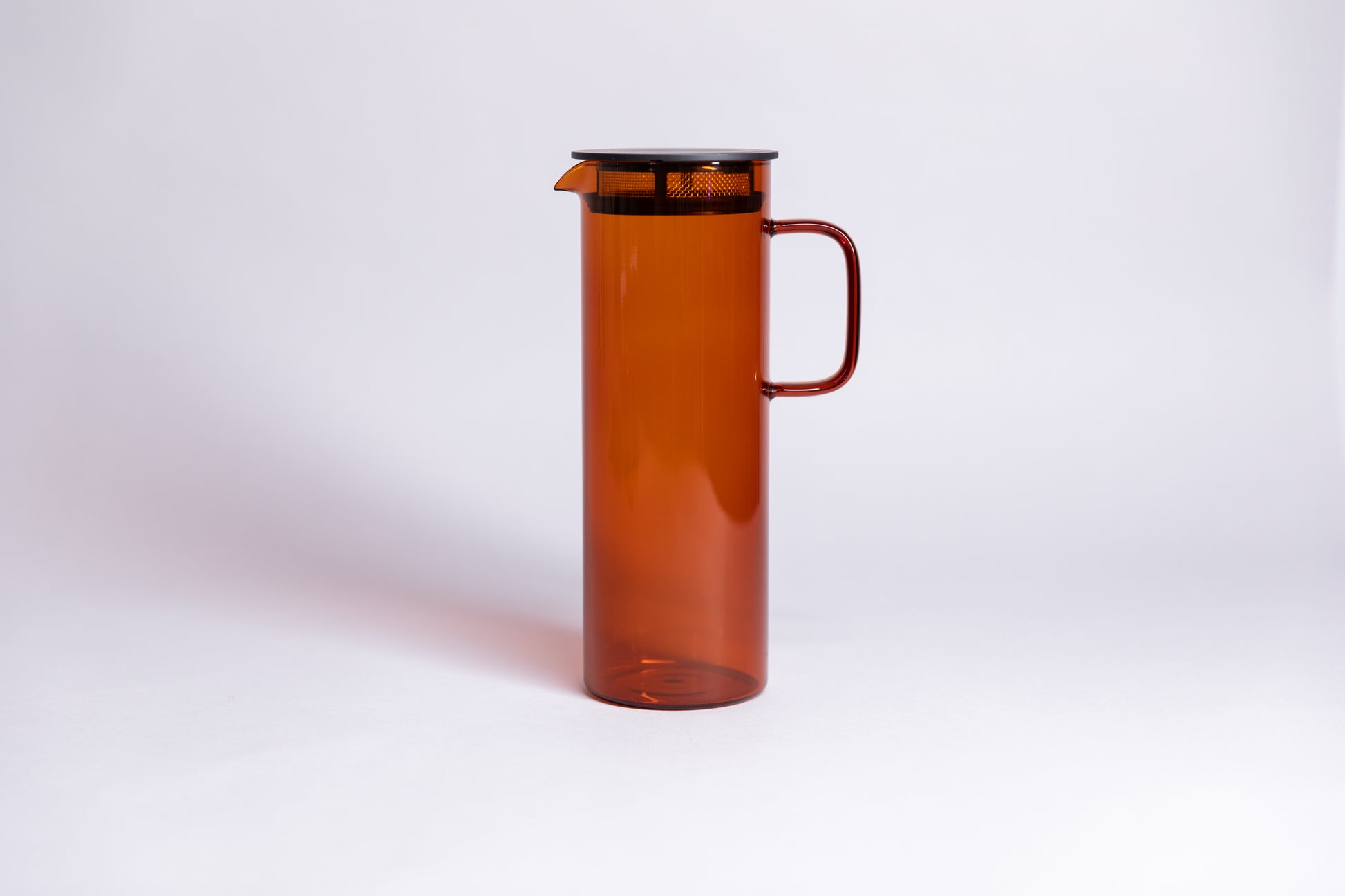 Cylindrical transparent amber colored glass pitcher with a fluted spout and rectangular handle with a black colored filtered lid. set on a a white background.