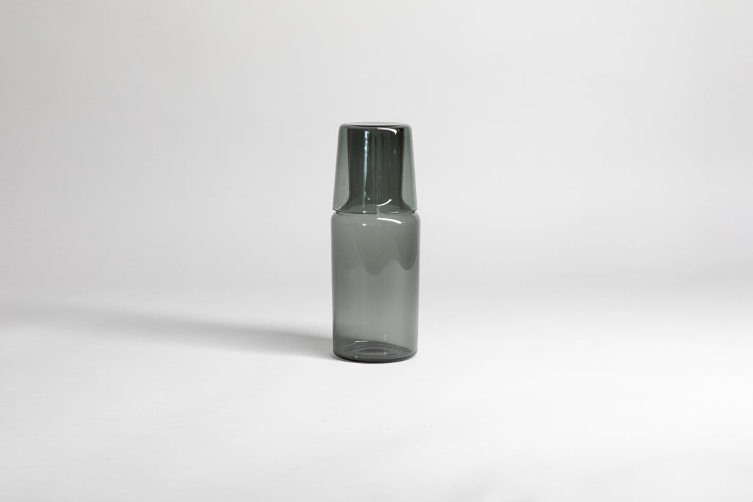 Transparent gray carafe with matching glass cup flipped upside down on carafe top of carafe. Set on a light gray background
