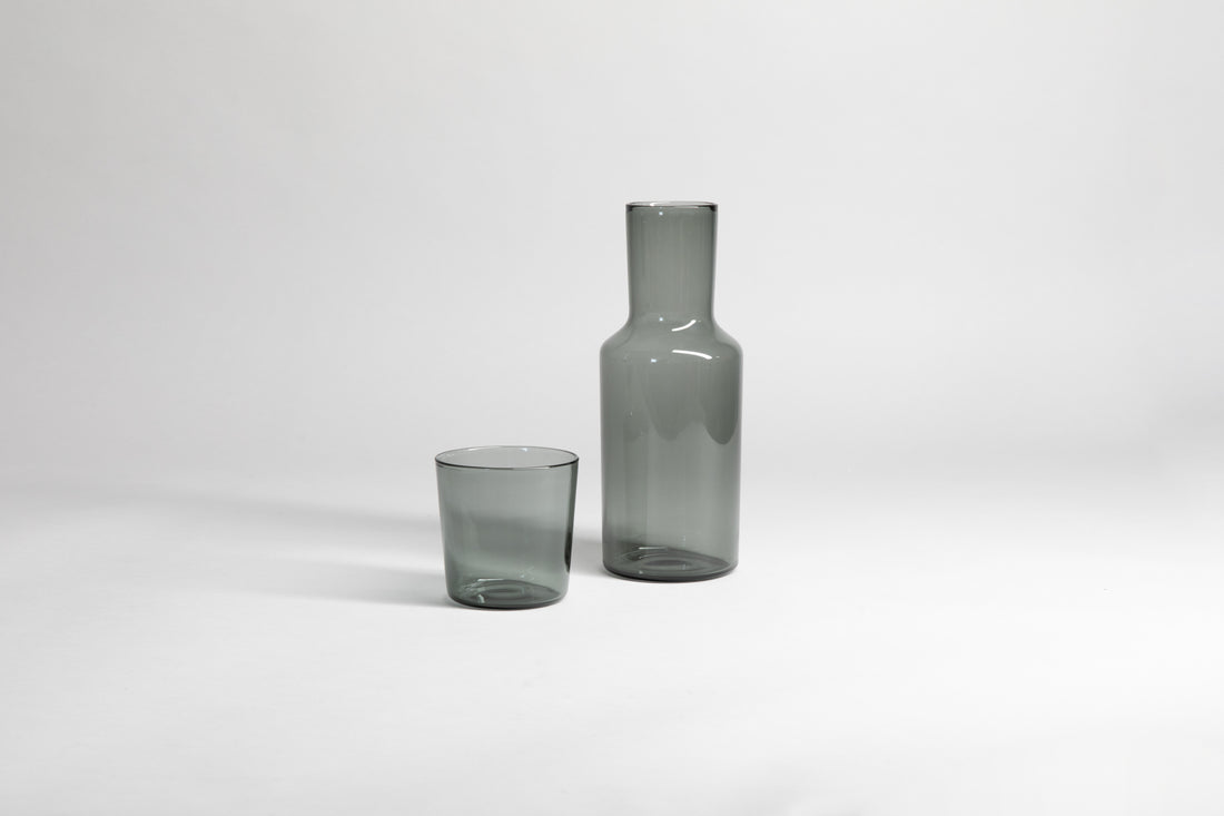 Transparent gray glass carafe with matching glass cup set on the side. Set on a light gray background