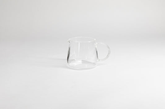 Round glass server with glass handle and fluted spout. On a light grey background.