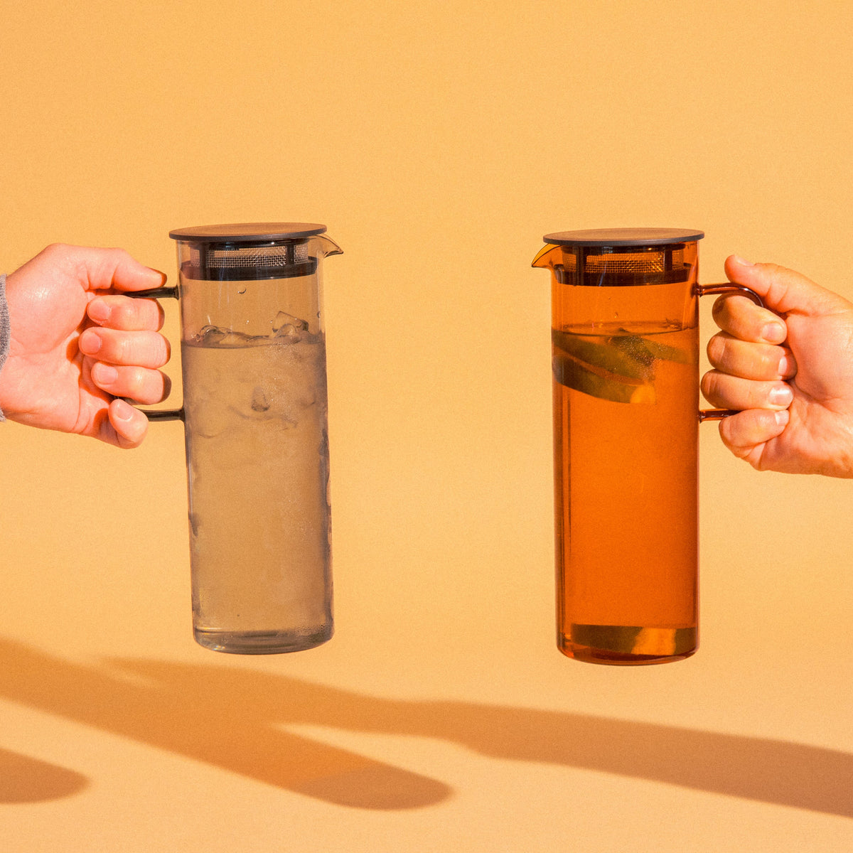 Two cylindrical transparent one gray color glass and one amber colored pitcher with a fluted spout and rectangular handle with a black colored filtered lid. set on a a light orange background background. Held by 2 different hand