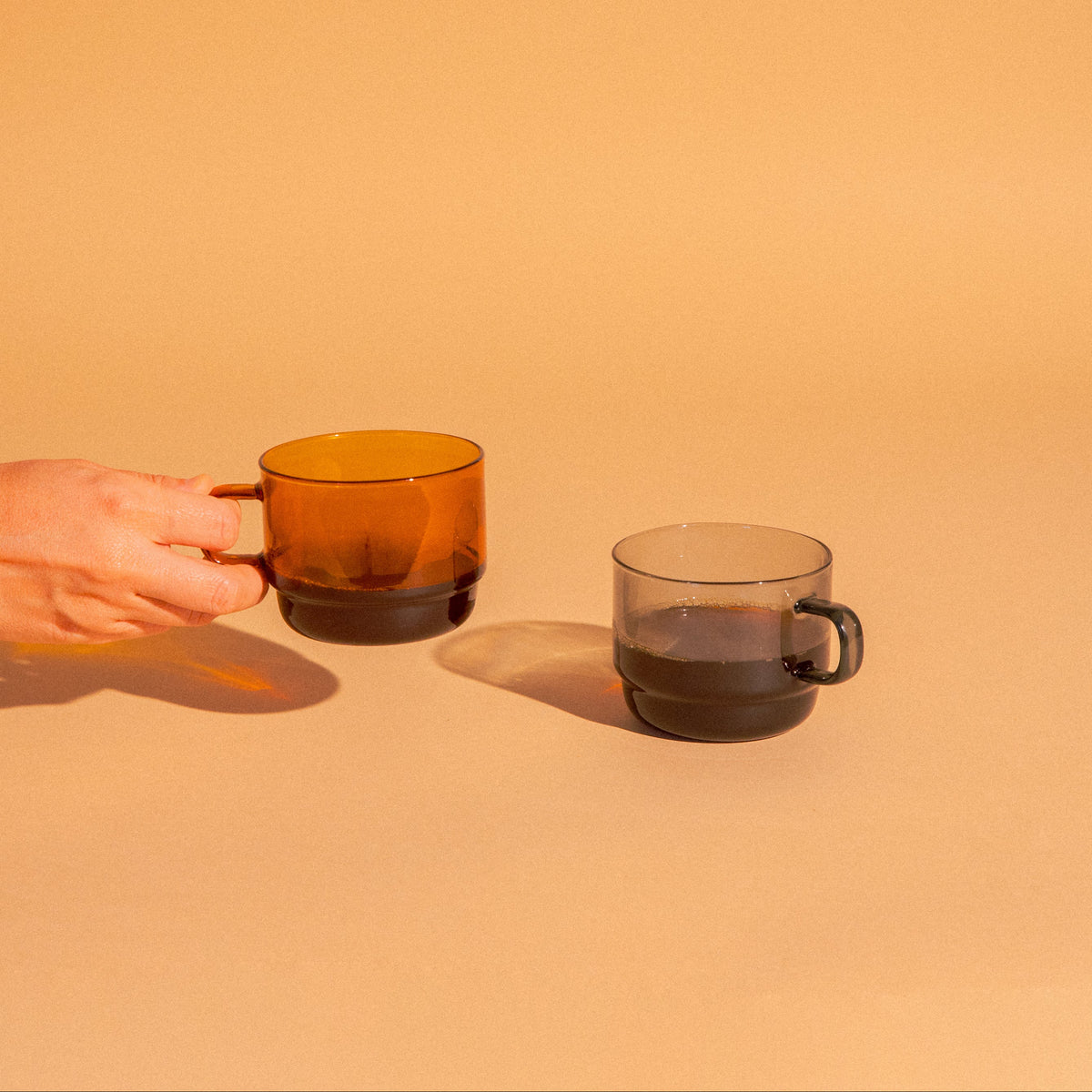 Hand holding amber glass mug with coffee. Transparent gray glass mug with coffee set on a light terracotta background