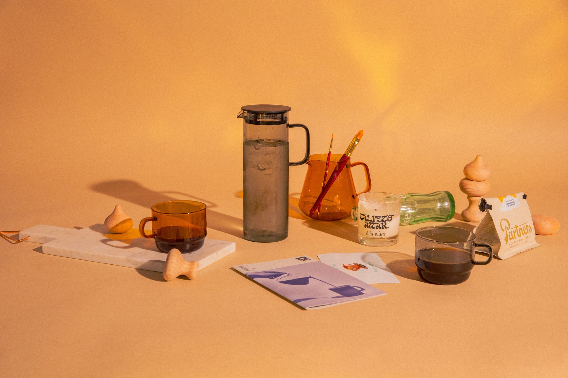 Transparent glass amber mug filled with coffee and transparent glass amber tea/coffee server with 2 decorative paint brushes. Transpartment cylindrical grey pitcher with handle filled with water. Transparent grey glass mug with coffee. Objects surrounded with decorative art pieces. set on a orange background