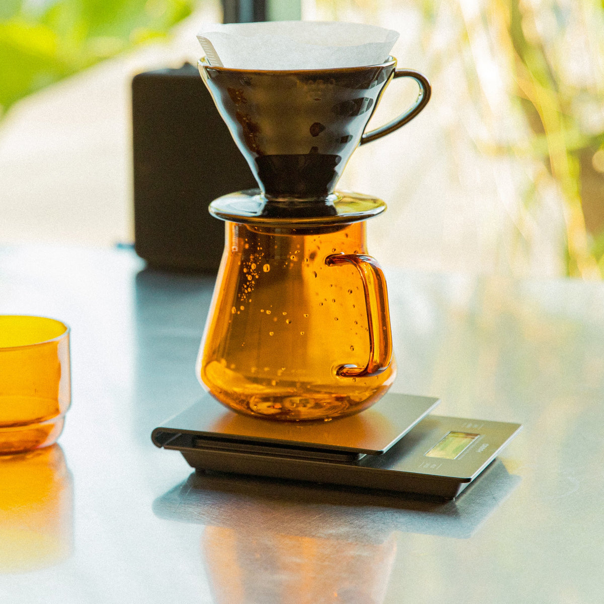 gray and amber colored transparent glass mugs. Black HARIO V60 top of amber glass transparent server with handle sitting on a black pour over scale.
