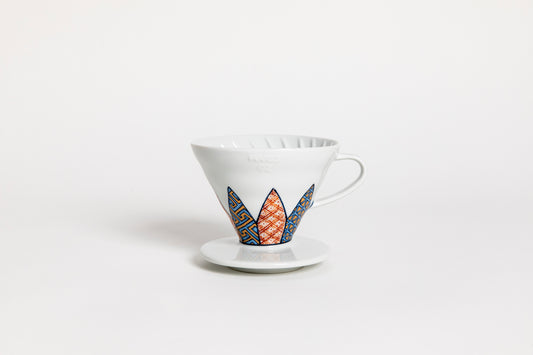 White cone-shaped ceramic coffee dripper with handle and round base, featuring Shonzui (祥瑞) pattern with auspicious symbols like pine, bamboo, and plum blossoms on a geometric tortoise-shell design. set on a white backaground.