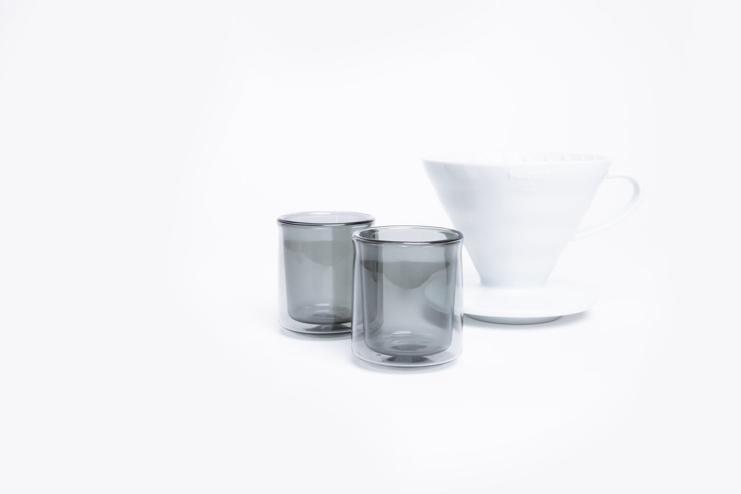 Two smoked glass tumblers and a white cone dripper with handle on a white background