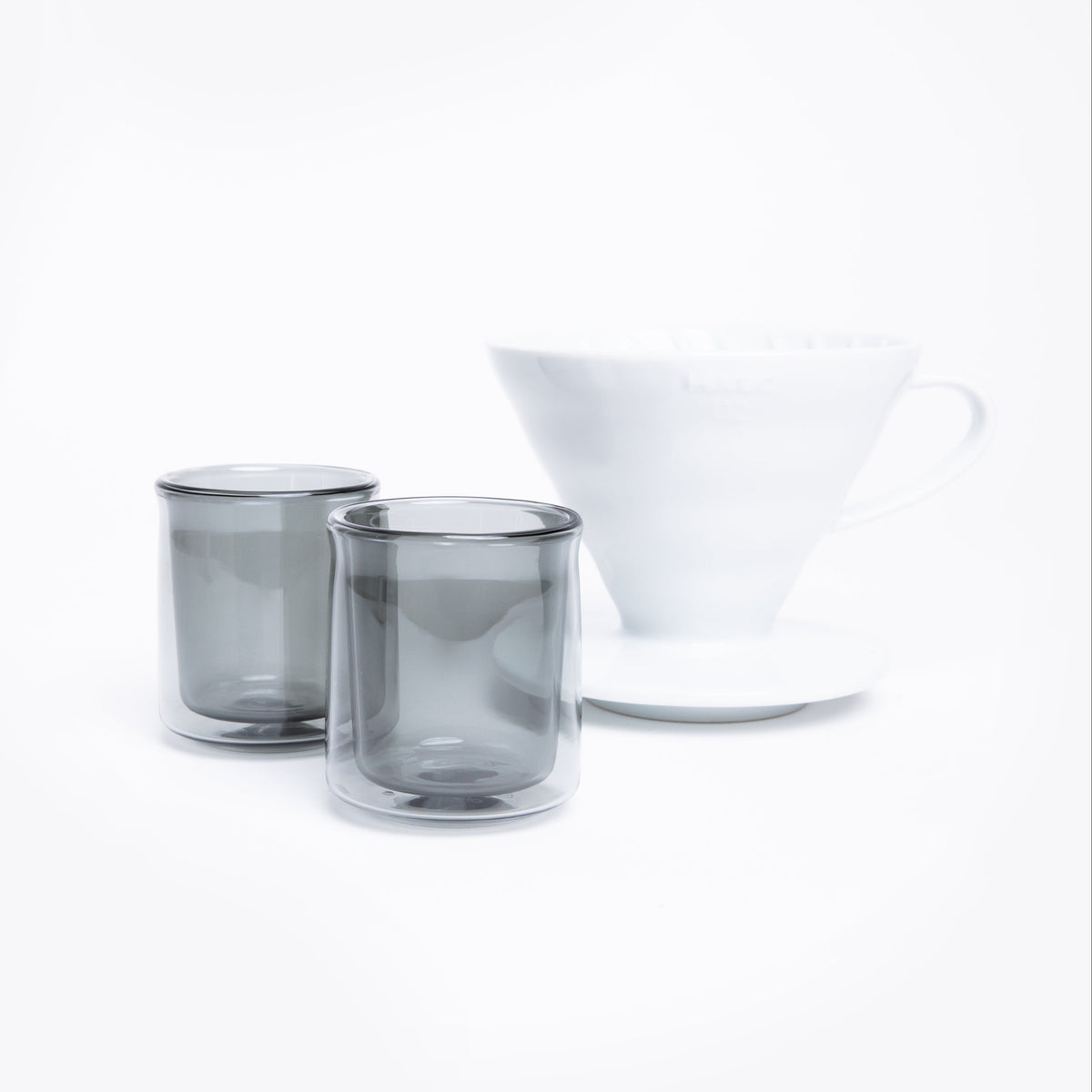 Two smoked glass tumblers and a white cone dripper with handle on a white background