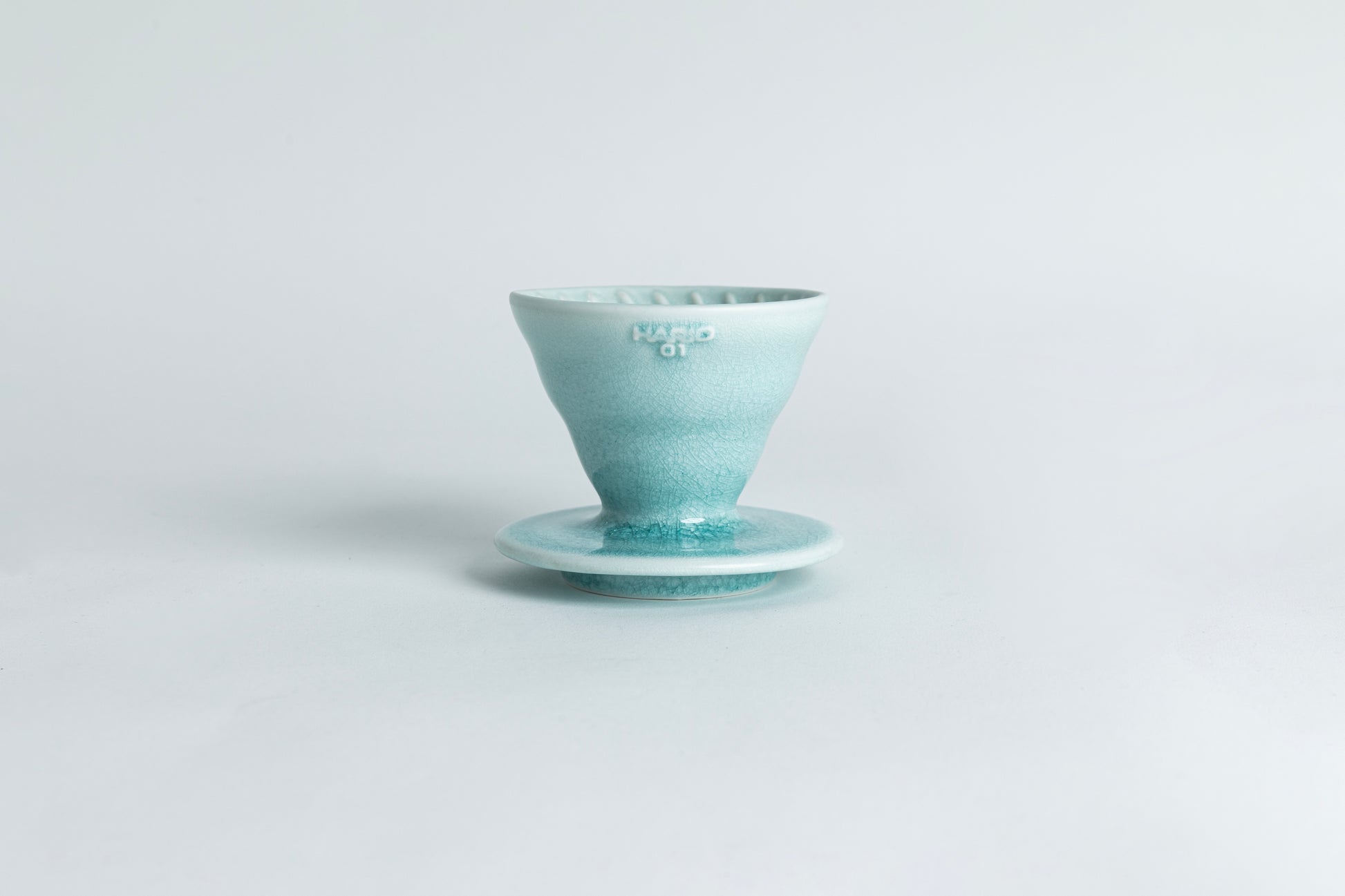 A ceramic pour-over cone with a saucer base featuring a gradient crackle glaze from light teal to teal. The text 'HARIO 01' is displayed on the cone. Set against a light gray background.