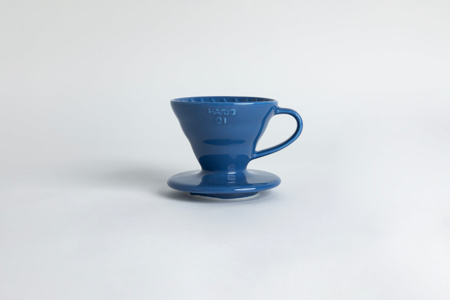 Blue ceramic cone shaped ceramic coffee dripper with handle and round base. Spiral ribbed on the inside cone, with the HARIO 01 logo on the side. Set on a light gray
background