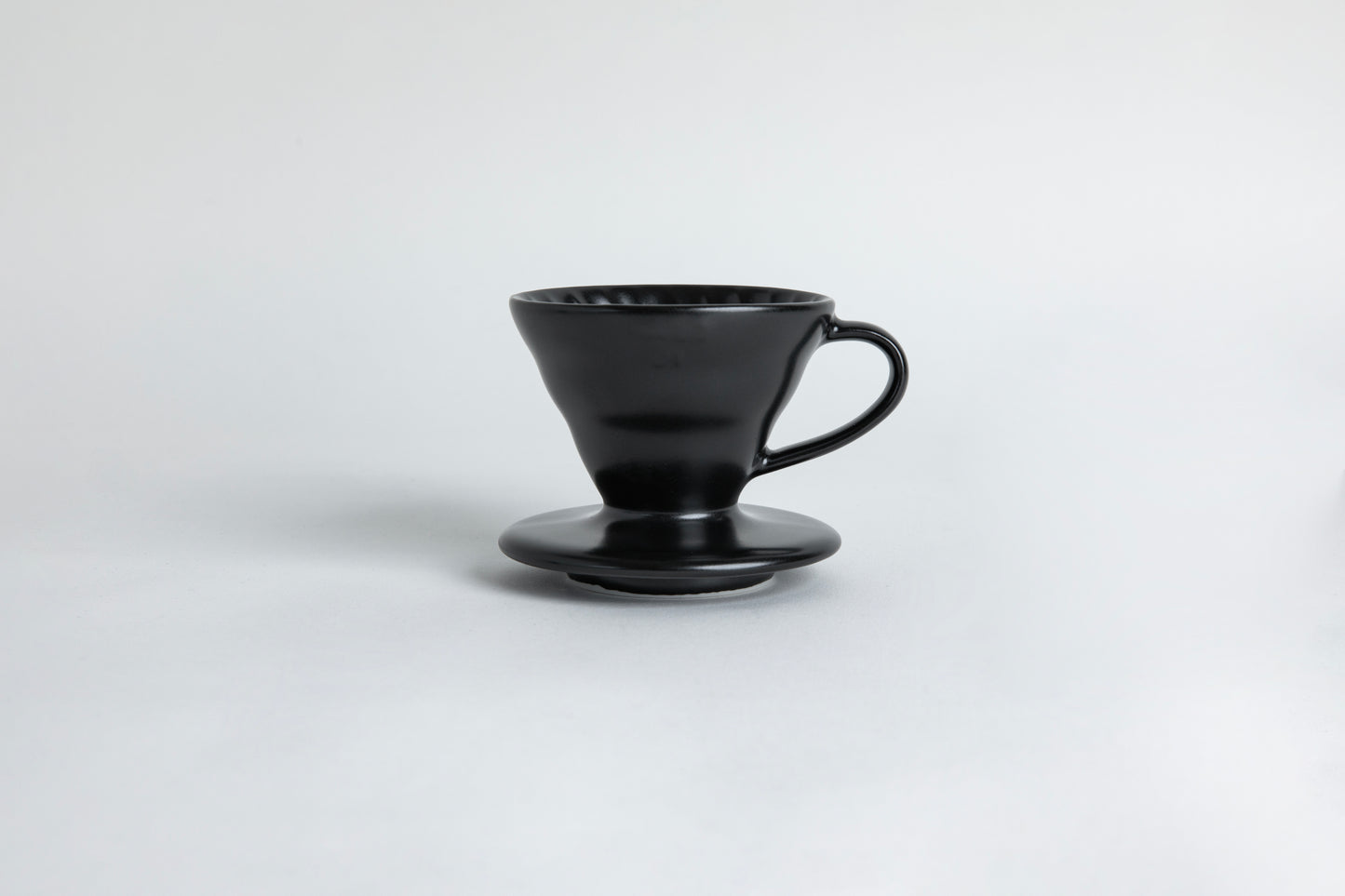 matte black cone shaped ceramic coffee dripper with handle and round base. Spiral ribbed on the inside cone. Set on light gray background