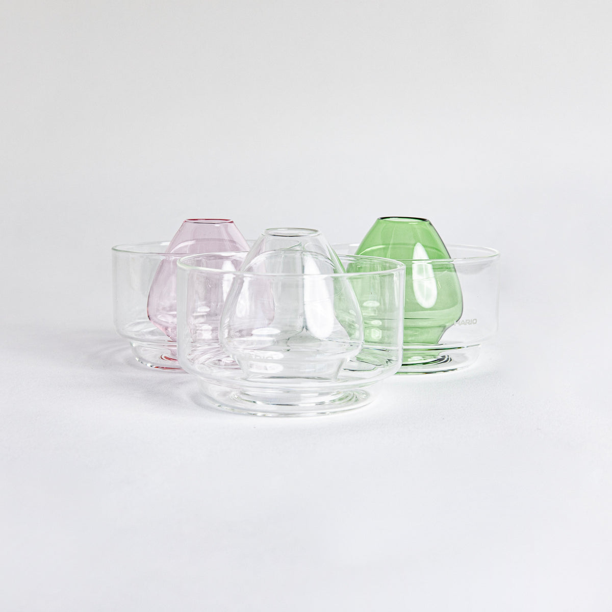 Three side by side images of a dome-shaped in the colors clear, pink and green clear glass matcha whisk holder with a smooth, tapered form, placed inside a clear glass base. Displayed on a white background with 'Hario' branding