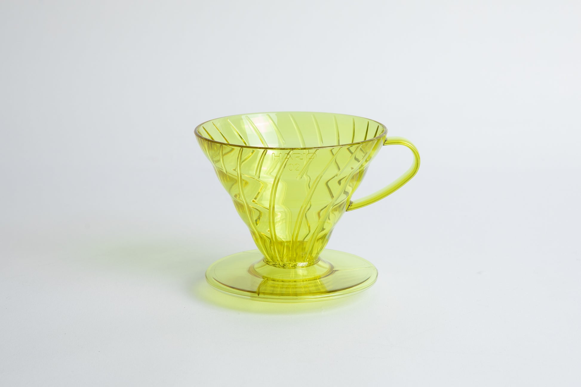 set on it's side the Size 02 Plastic V60 Dripper in the color transparent Chartreuse (Aurora) with a cylindrical base and curved handle. Set on a white background