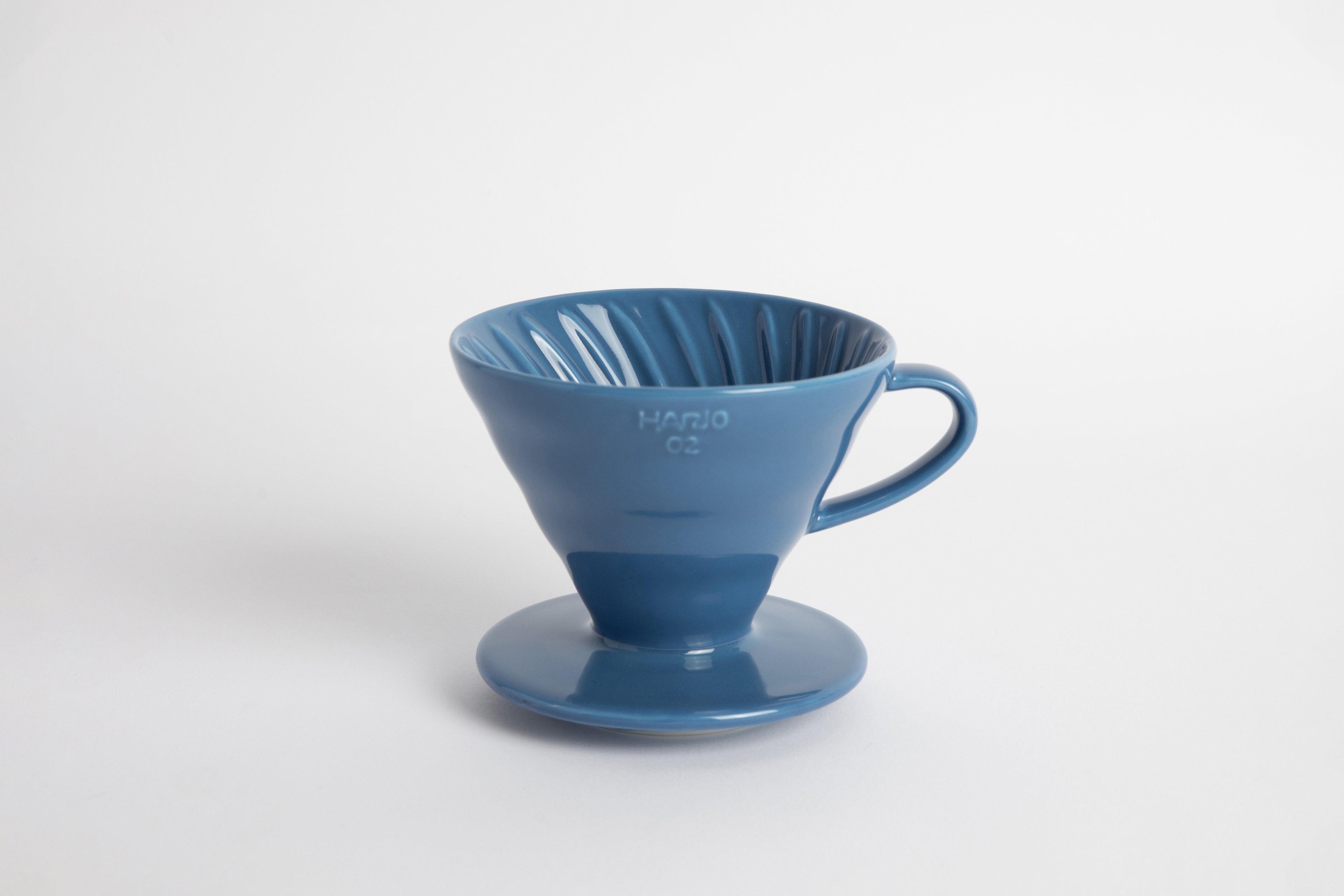 Blue colored 60 degree cone shaped ceramic coffee dripper with handle and round base. Spiral ribbed on the inside cone. Set on white background