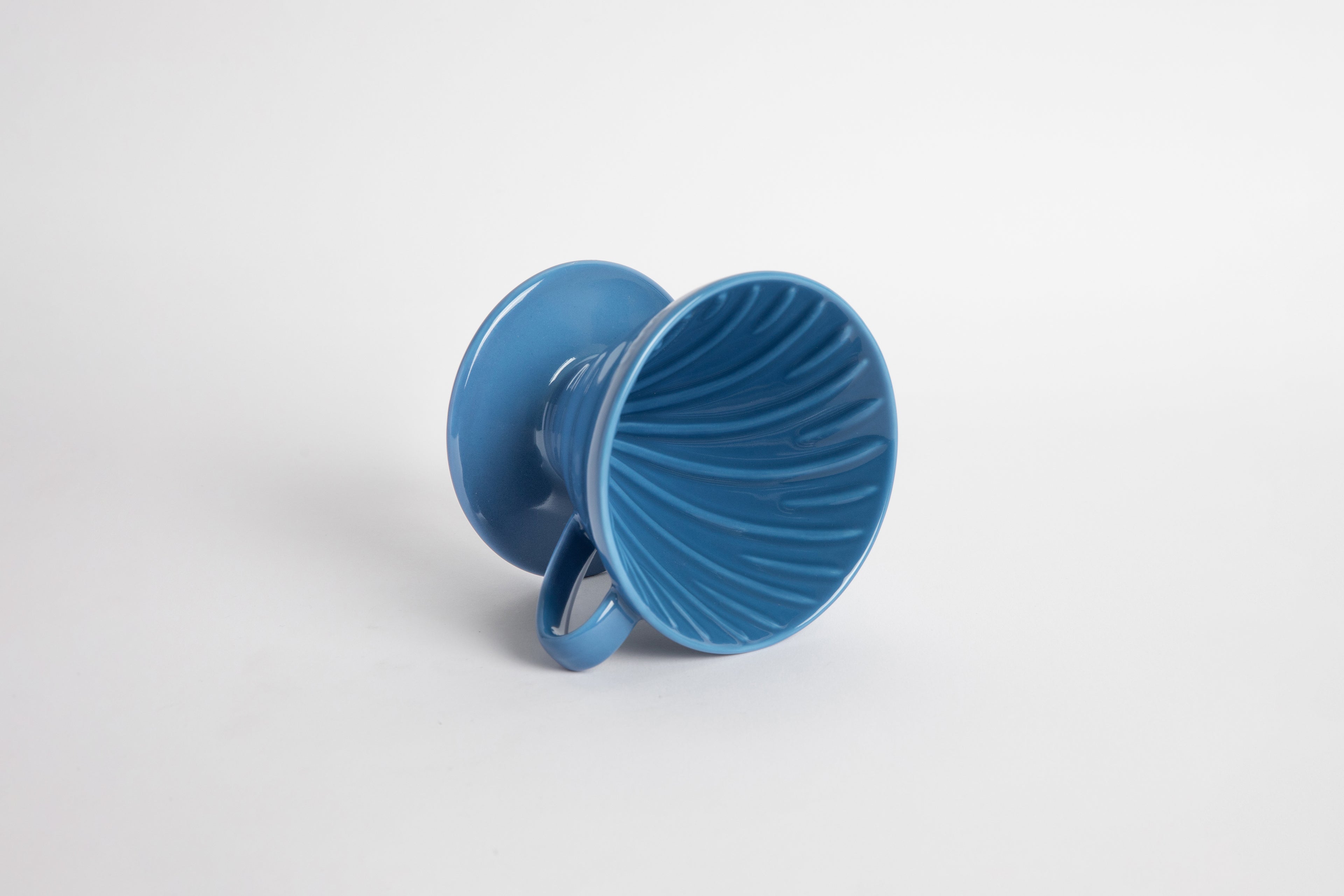 Blue colored 60 degree cone shaped ceramic coffee dripper with handle and round base. Spiral ribbed on the inside cone. Set on white background