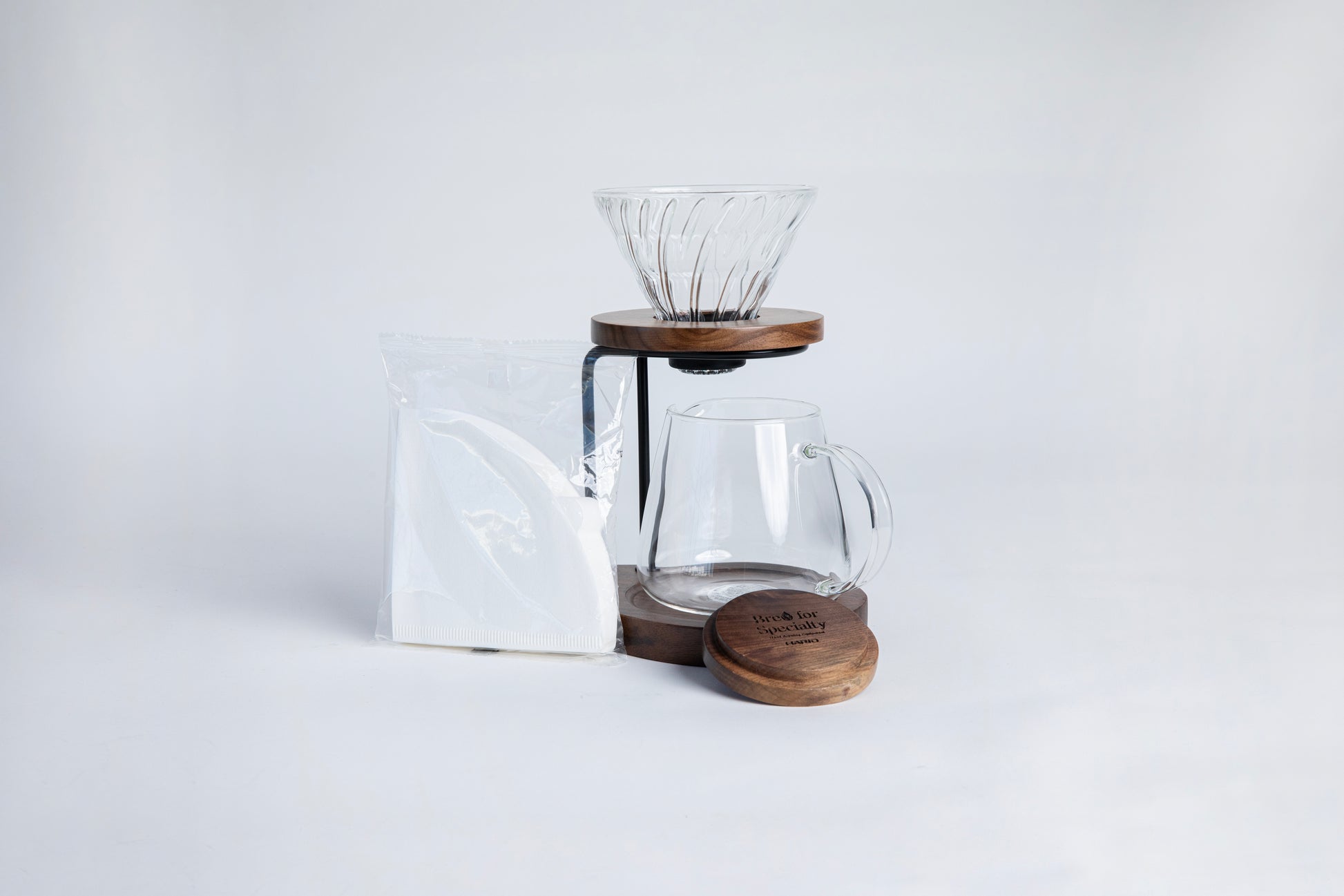 Clear all glass cone shaped coffee dripper with ribs, sitting in a round walnut wood base. Dripper is on a on metal and walnut wood hanging stand underneath a a tulip shape glass decanter with glass handle. With a walnut lid for the decanter and a triangular shaped filters in a cellophane bag. set on a white background.