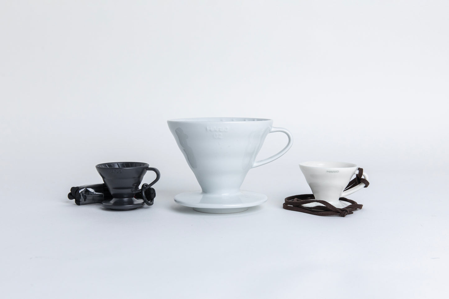 Three cone-shaped coffee drippers with handles and round bases, arranged side by side on a white background. The first is a mini matte black dripper with a brown rope tied around the handle. Next to it is a standard 02-size ceramic dripper in a classic finish, and beside that, a mini white ceramic dripper with the same cone shape and design. set one a white background.