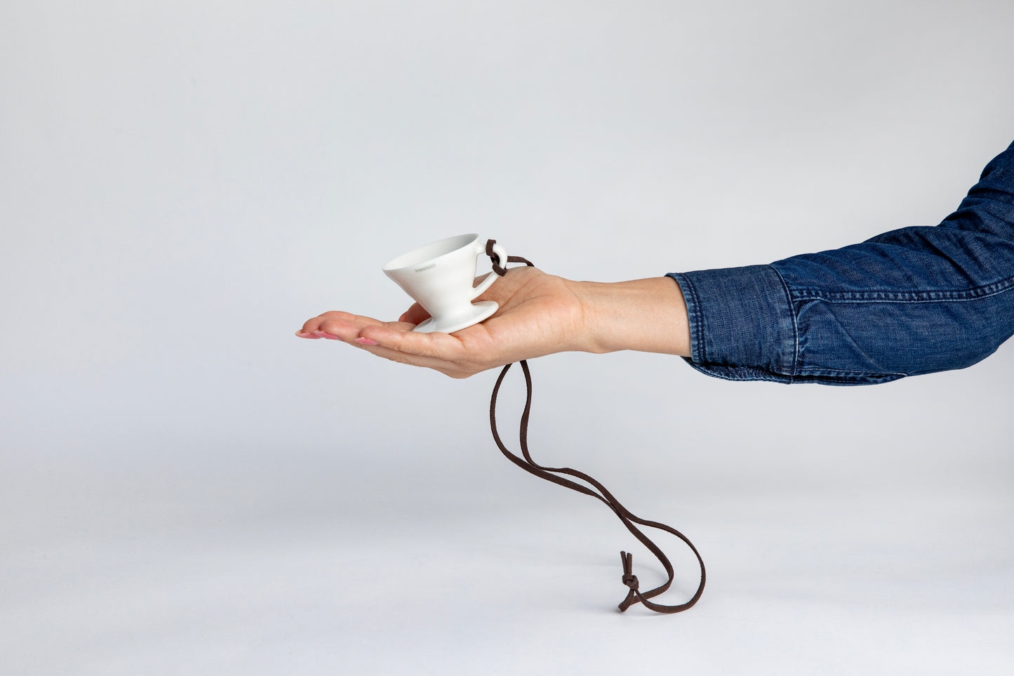 hand holding the white black cone shaped ceramic coffee dripper cup with handle and round base. with a brown rope tied on the handle, showcasing the size of the small cup can fit in the palm of a  hand. Set on white background