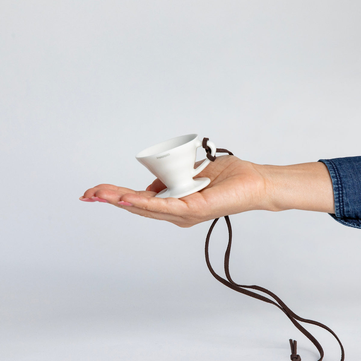 hand holding the white black cone shaped ceramic coffee dripper cup with handle and round base. with a brown rope tied on the handle, showcasing the size of the small cup can fit in the palm of a hand. Set on white background