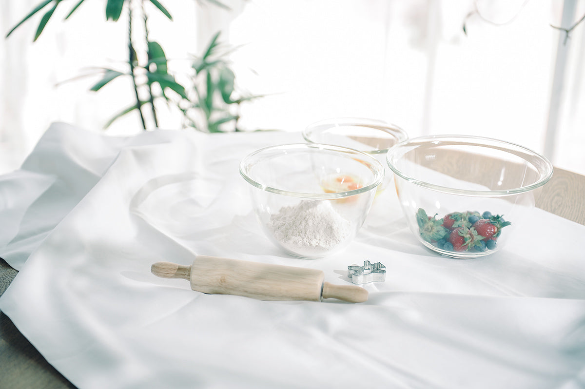 Three glass mixing bowls from small, medium, to large on a white linent table cloth crumpled across a round wood table with eggs in the small bowl, flour in the medium bowl, strawberries and blueberries in the large bowl, and next to a wood rolling pin and silver tin cookie cutter. Two green fronds in the background beside a white curtain.