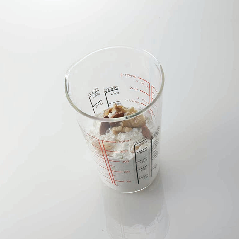 Tall glass measuring cup with small spout and red and black measurements on the inside and outside of the cup with flour and nuts inside on a white reflective background.