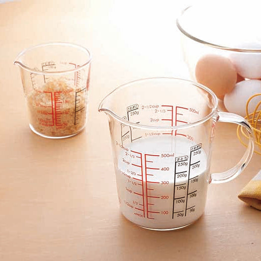 Glass measuring cup with spout, full glass handle, measurements in red and black on the inside and out, and filled with milk on a table setting with a small glass measuring cup with measurments in red and black on the inside and outside of the glass with bread crumbs inside. A yellow whisk on a folded yellow, white and beige checkered hand towel and a glass mixing bowl filled with brown and white eggs.