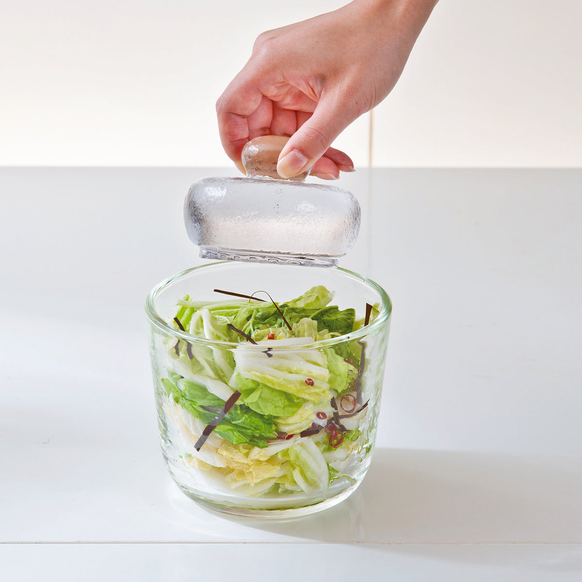 Woman's hand holding the knob of weighted glass stone above glass salting bowl with salad inside.