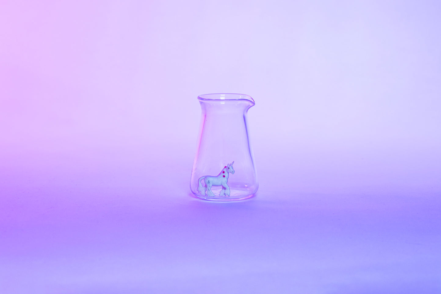 Conical glass flask-shaped coffee pitcher with small white unicorn toy inside and set against a light pink and purple background.