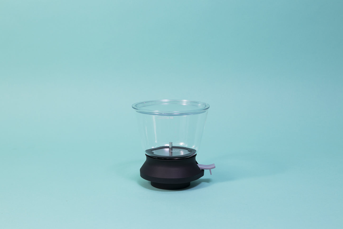 Cylindrical glass tea brewer and metal mesh strainer with clear plastic lid atop a black rubber base with plastic lever