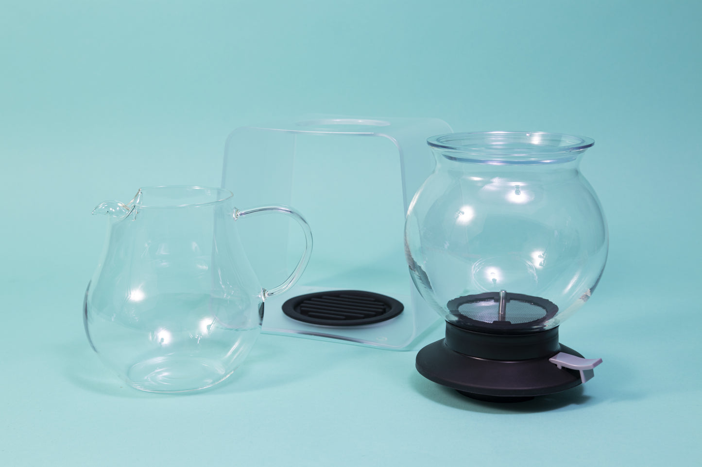 Globulous glass tear brewer and metal mesh filter with clear plastic lid atop a black rubber base with plastic lever switch next to a clear acrylic stand above an all glass bulbous server with handle