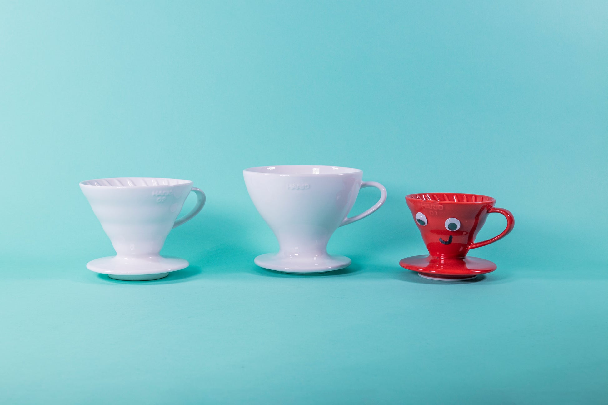 White ceramic cone shaped dripper with round base, wider and taller white ceramic cone shaped dripper with rounded edges and base, and a small red ceramic cone shaped dripper with round base and plastic eyes and a smiley mouth drawn on.