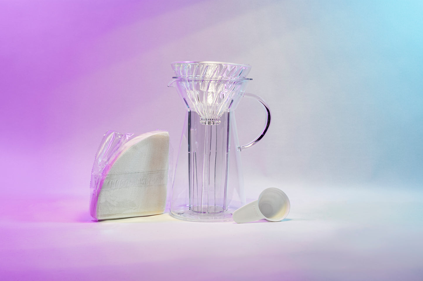 White cone shaped paper filters in a clear plastic bag next to a tapered glass dripper with fluted spout and all glass handle with a cone shaped glass dripper with ribs seated inside, and a white plastic coffee measuring spoon.