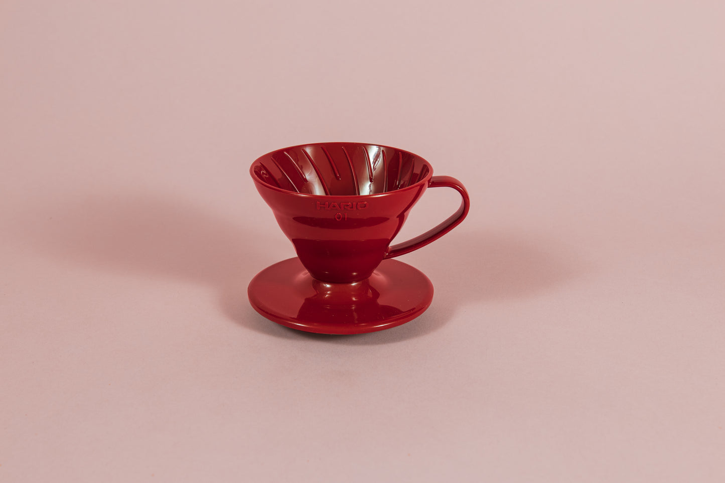 Red all plastic cone shaped dripper with handle and round base on a pink backdrop.