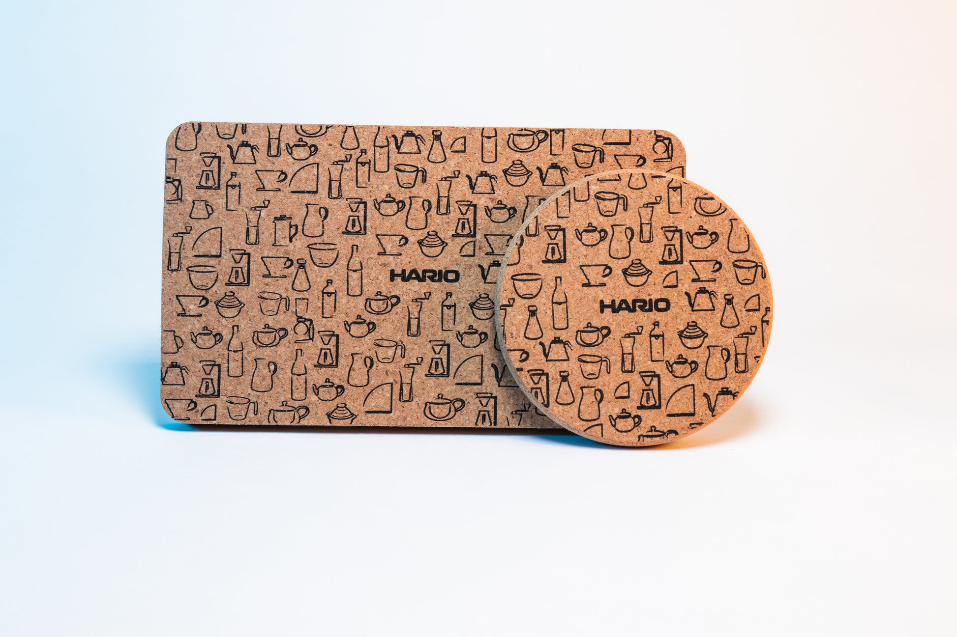 Rectangular cork coaster mat with black Hario logo and black pattern design of Hario products behind a circular cork coaster with black Hario logo and black pattern design of Hario products.