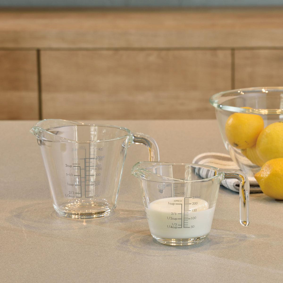One and two cup sized thick glass measuring cups with glass handles and measurements on both the outside and inside of the glass on a table next to a glass mixing bowl with lemons inside and seated on a hand towel. Beside the towel is another lemon and the one cup measuring glass is filled two-thirds of the way with milk.