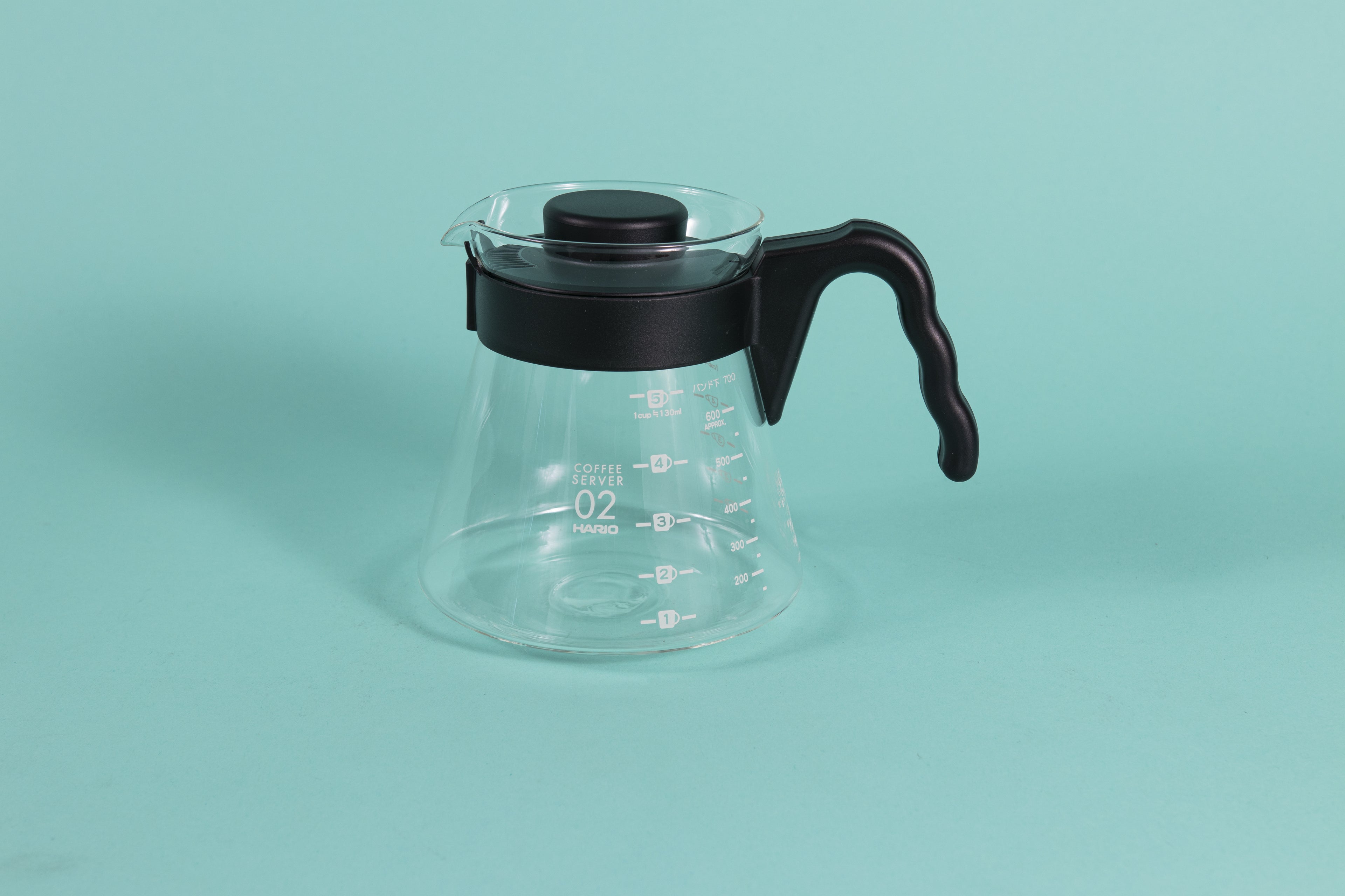 Glass coffee server with white text and level markings with black hard plastic handle and lid on a teal backdrop.