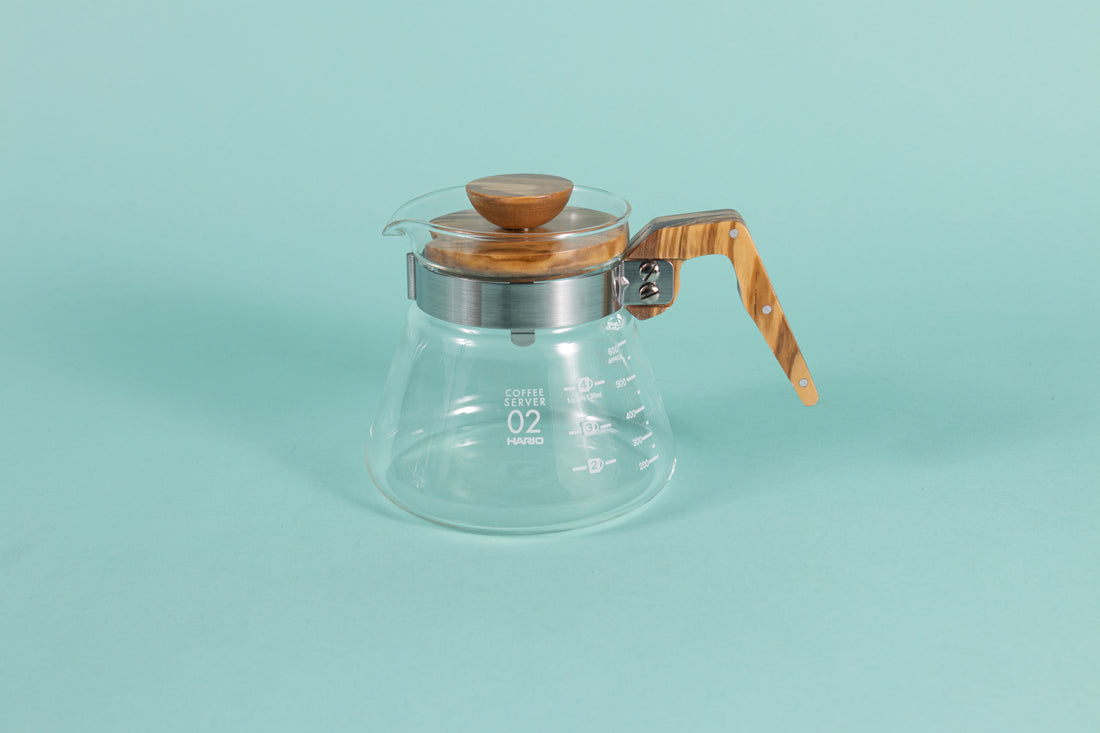 Glass coffee server with white text and markings with a brushed metal collar and wooden handle and lid on a teal backdrop.