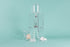 Tall clear acrylic tower stand with water reservoir and drip spout sitting over a glass cylinder with metal mesh filter for coffee grounds, with all glass coffee server with handle underneath and paper filter pack, server lid, and white plastic scoop on a teal background