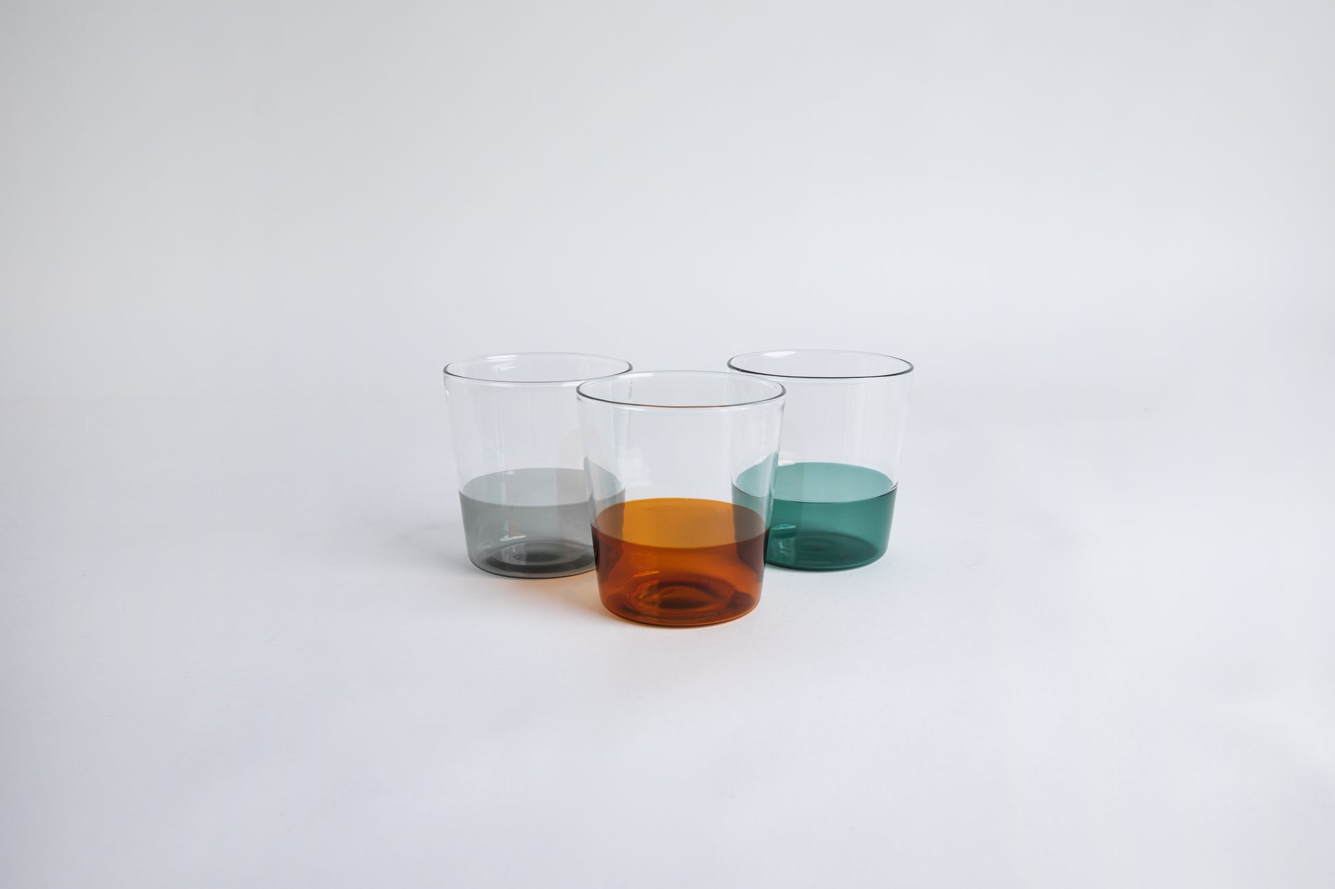 3 dual colored clear glass cup side by side, one cup top half is clear and second half colored amber, one cup top half is clear and second half colored gray, one cup top half is clear and second half colored green.  set on a white background