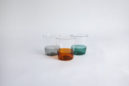 3 dual colored clear glass cup side by side, one cup top half is clear and second half colored amber, one cup top half is clear and second half colored gray, one cup top half is clear and second half colored green.  set on a white background