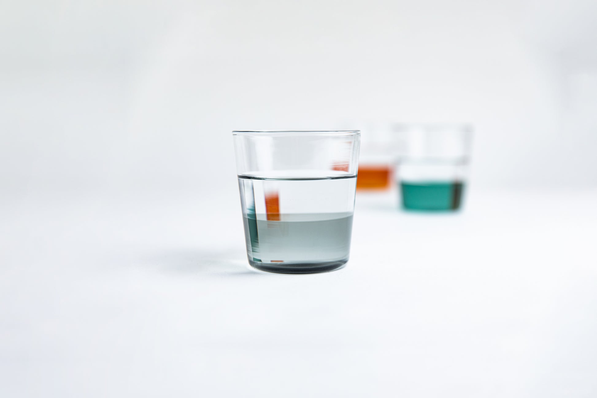Dual colored clear glass cup, top  half is clear and second half colored gray filled with water. Foreground blur photo of dual colored clear glass cup, top  half is clear and second half colored green and other cupnis amber set on a white background 