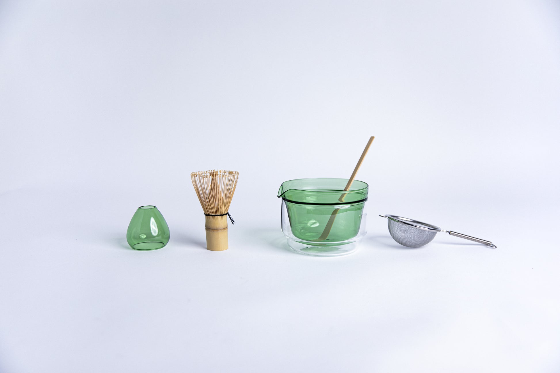 Matcha tea-making tools including a bamboo whisk, green glass dome shape bamboo whisk holder, Double-walled glass cup with green interior, stick tool and metal sieve. set on a white background.