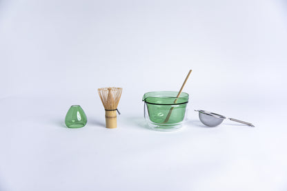 Matcha tea-making tools including a bamboo whisk, green glass dome shape bamboo whisk holder, Double-walled glass cup with green interior, stick tool and metal sieve. set on a white background.