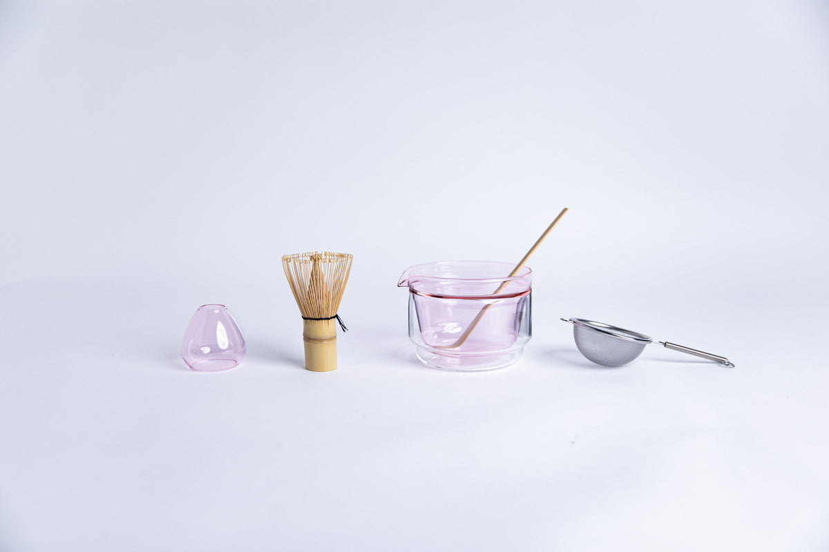 Matcha tea-making tools including a bamboo whisk, pink glass dome shape bamboo whisk holder, Double-walled glass cup with pink interior, stick tool and metal sieve. set on a white background.